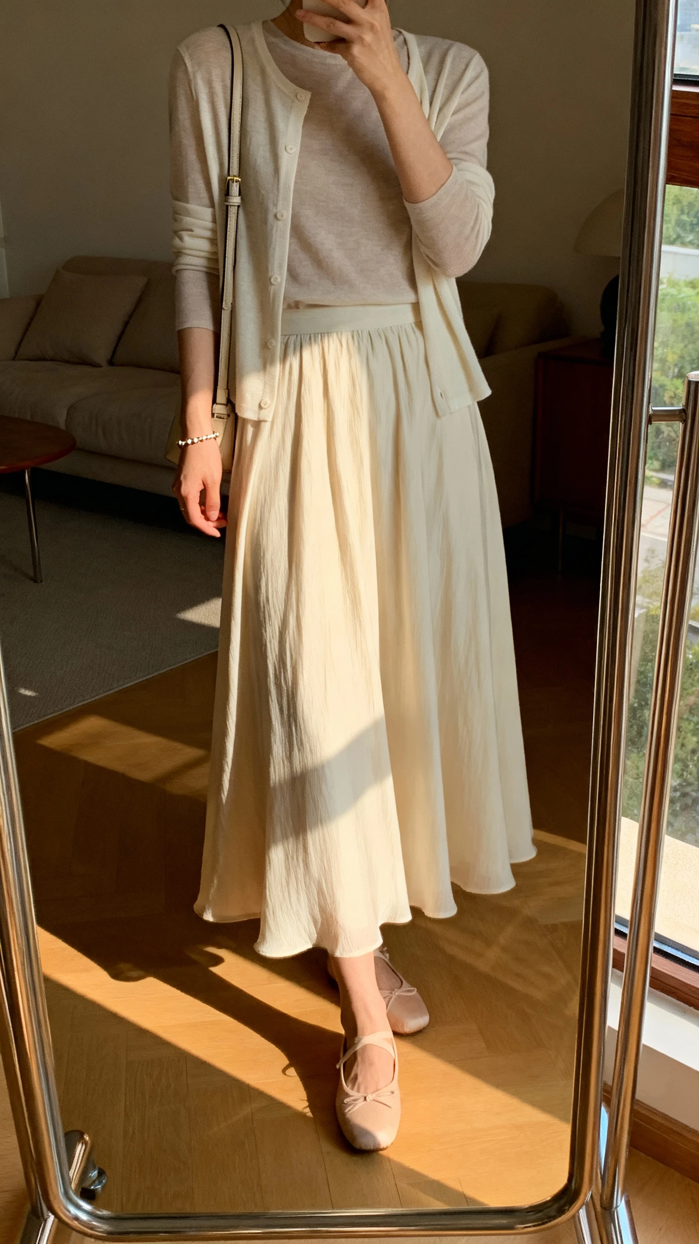 iPhone side-angle mirror selfie of a woman wearing a flowing A-line long skirt with a soft lightweight layer (cardigan or wrap) over a simple long-sleeve top, ballet flats, dainty bracelet, and a small shoulder bag, face not visible, living room full-length mirror, gentle afternoon window light, iPhone photo quality.