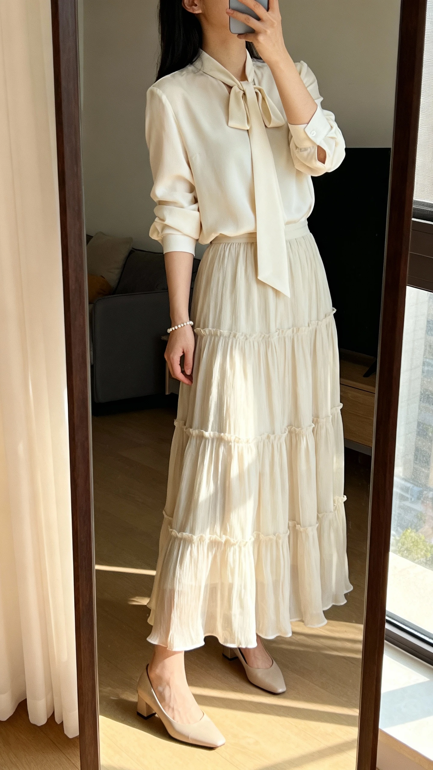 iPhone side-angle mirror selfie of a woman wearing a flowing tiered skirt with a soft tie-neck blouse, delicate bracelet and low heels, face not visible, cropped at neck, living room full-length mirror with gentle afternoon light, iPhone photo quality.