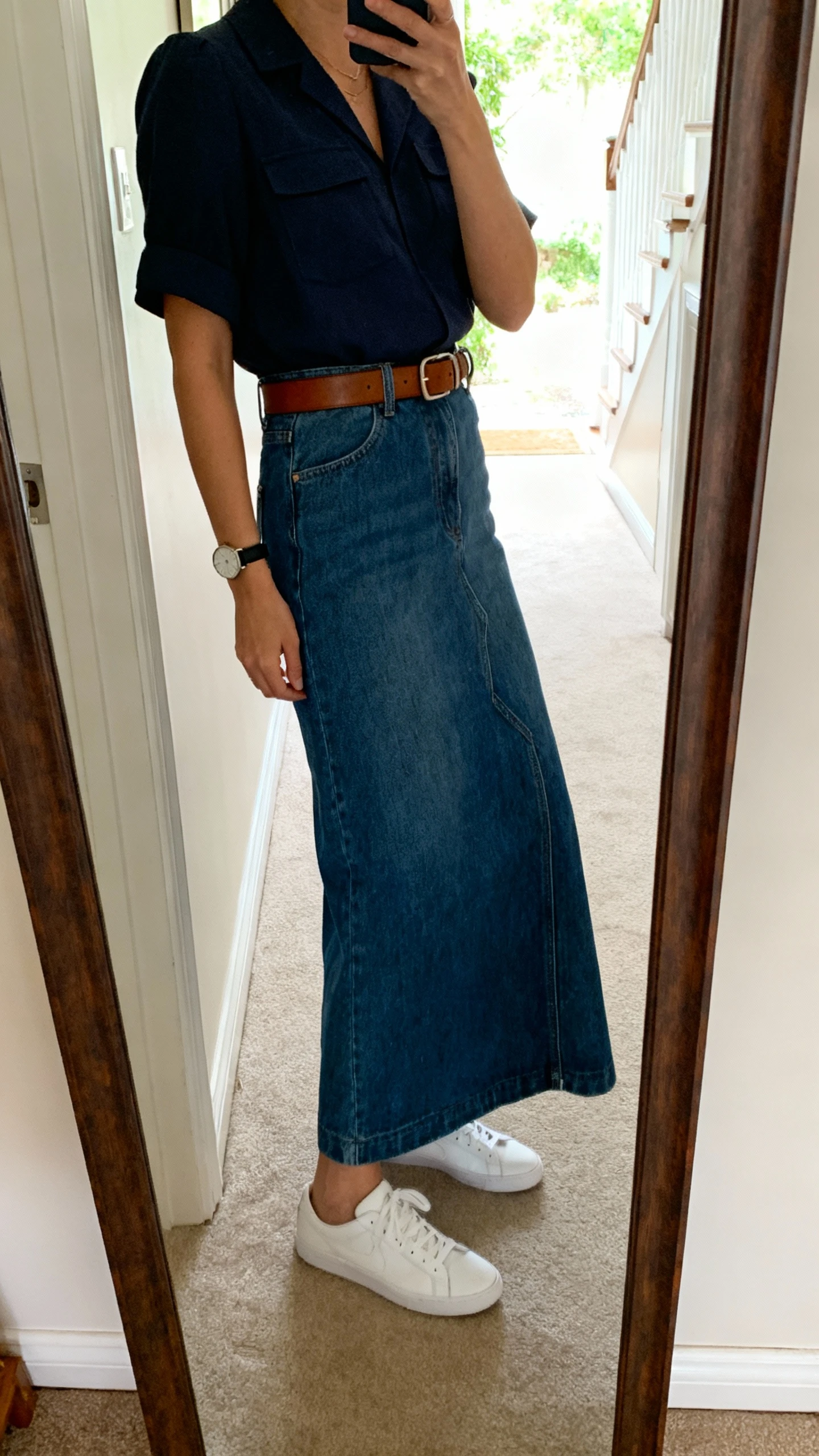 iPhone side-angle mirror selfie of a woman wearing a denim maxi skirt with a structured top, leather belt, clean white sneakers, and a simple watch, face not visible, entryway full-length mirror with natural daylight, iPhone photo quality.