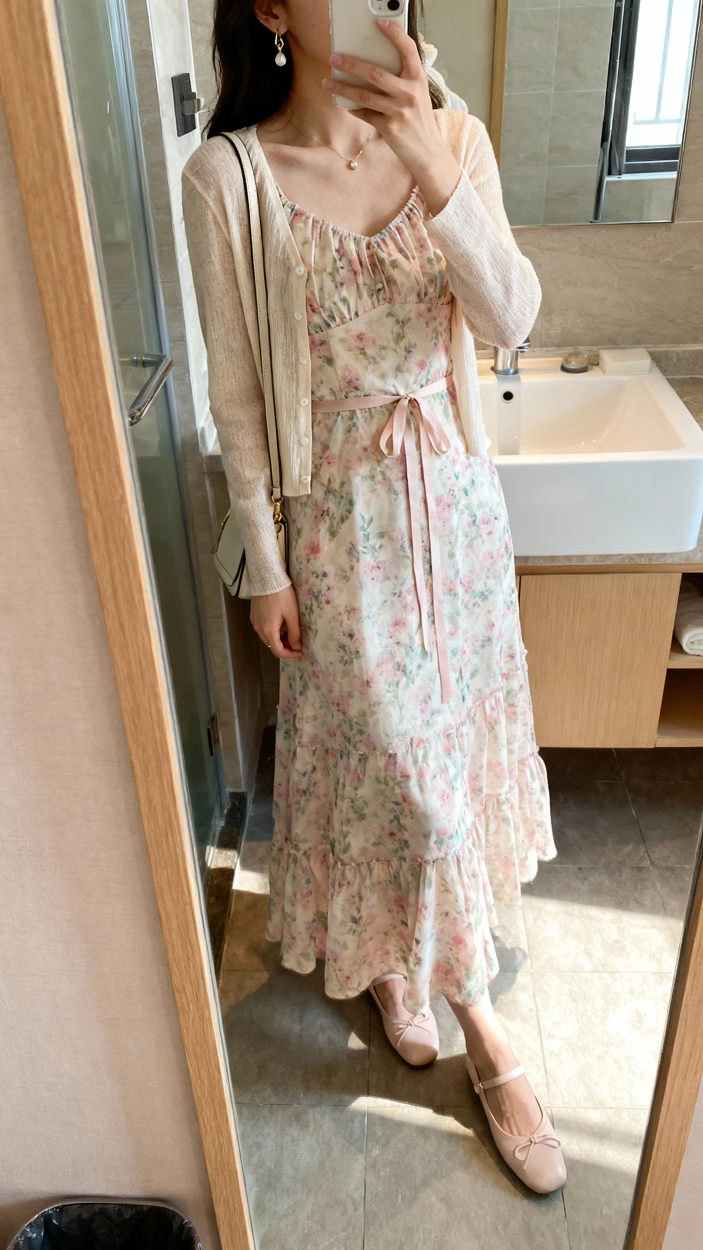 iPhone side-angle mirror selfie of a woman wearing a romantic pastel floral maxi dress with a lightweight cardigan and ribbon-tie details at the waist, dainty earrings, ballet flats, and a small shoulder bag, face not visible, bathroom mirror, gentle morning light, iPhone photo quality.