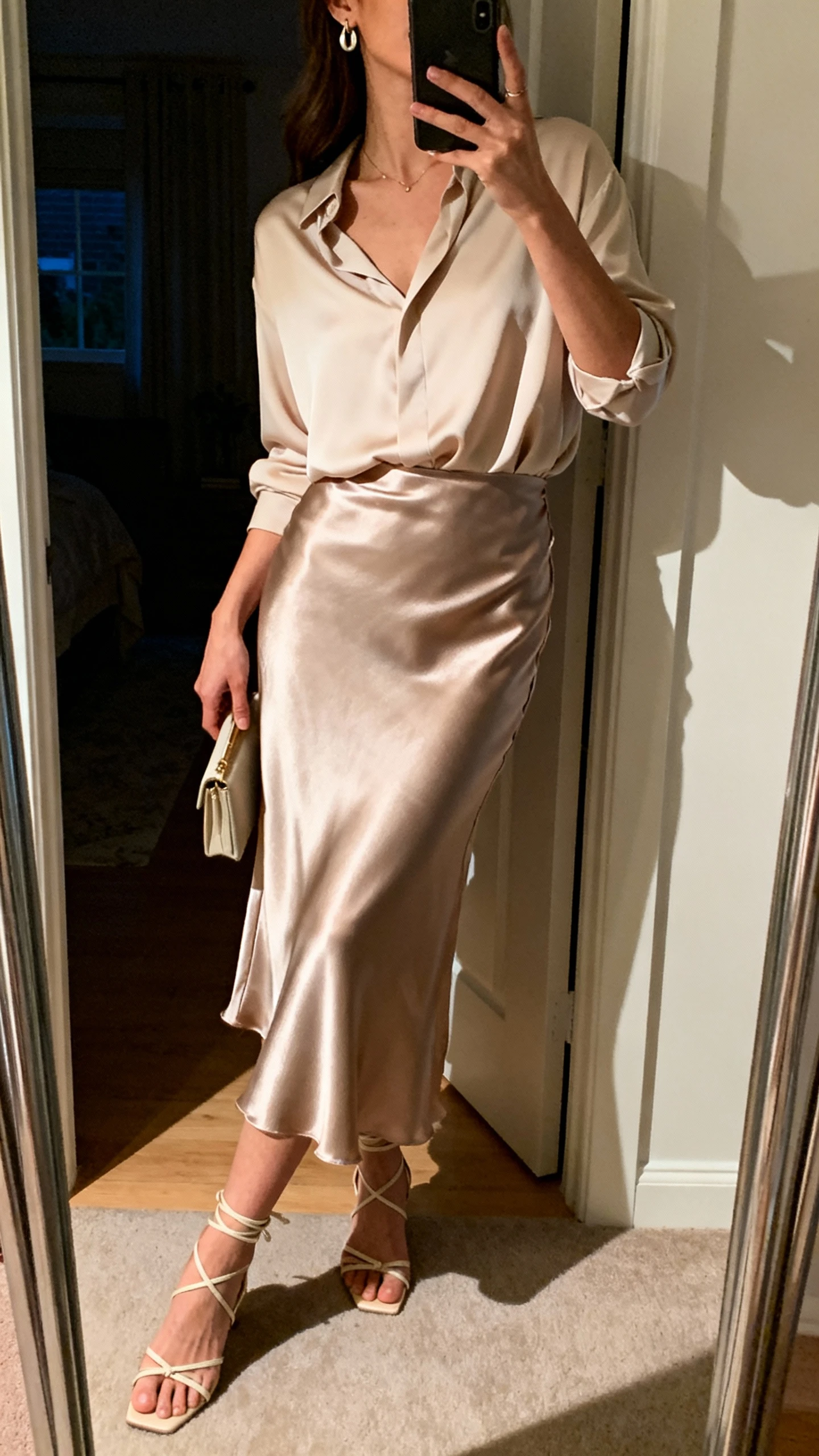 iPhone side-angle mirror selfie of a woman wearing a satin slip-style maxi skirt, soft drapey blouse tucked in, delicate earrings, strappy low heels, and a small clutch, face not visible, full-length mirror, evening indoor lighting, iPhone photo quality.