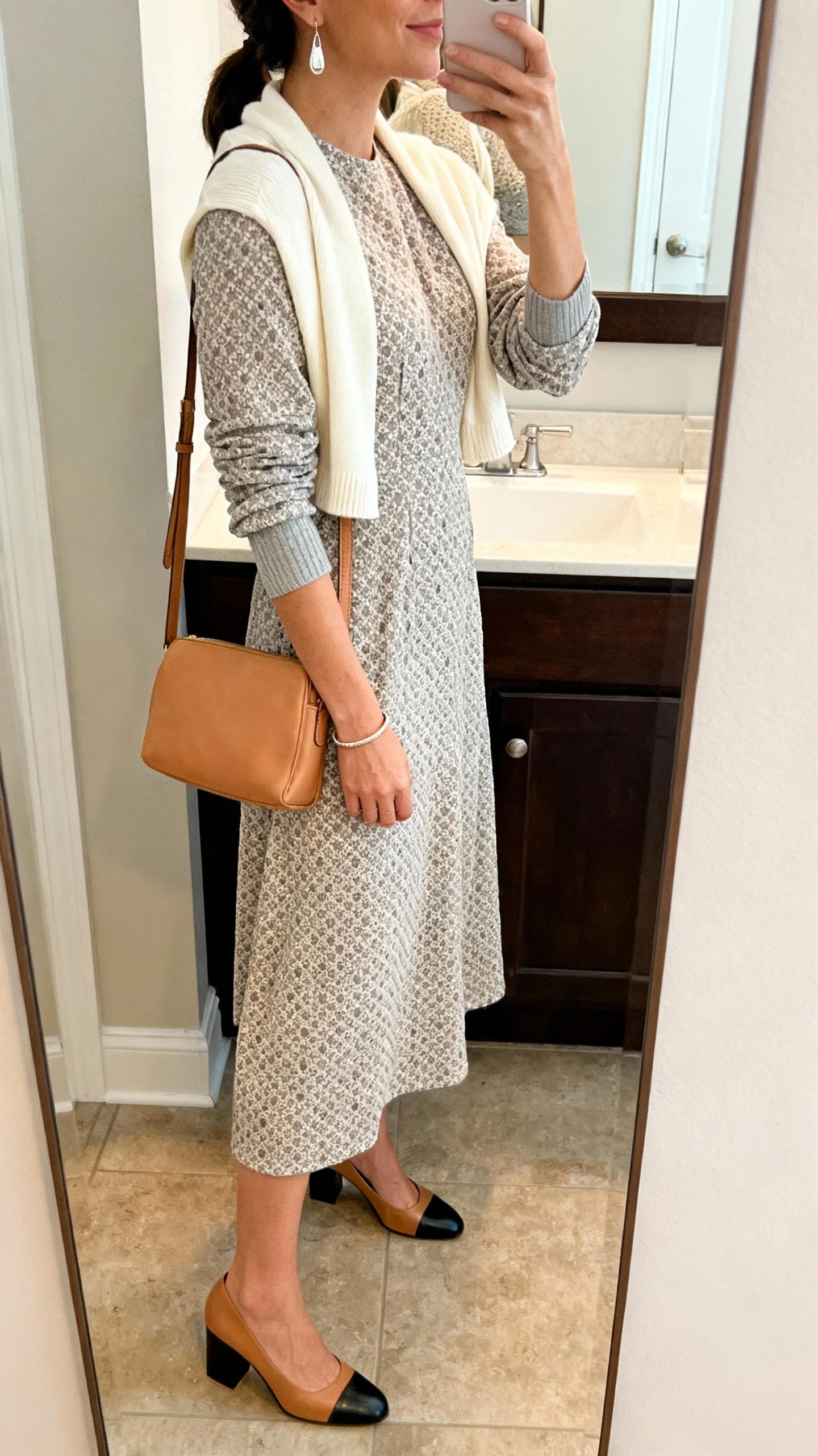 iPhone side-angle mirror selfie of a woman wearing a Swiss dot or tone-on-tone jacquard midi dress with elbow sleeves, a thin knit shrug or cropped cardigan in cream or soft grey, heeled Mary Janes or block-heel pumps in tan or black, delicate drop earrings, a simple bangle, and a camel leather shoulder bag; face not visible, bathroom mirror, warm indoor lighting, iPhone photo quality.