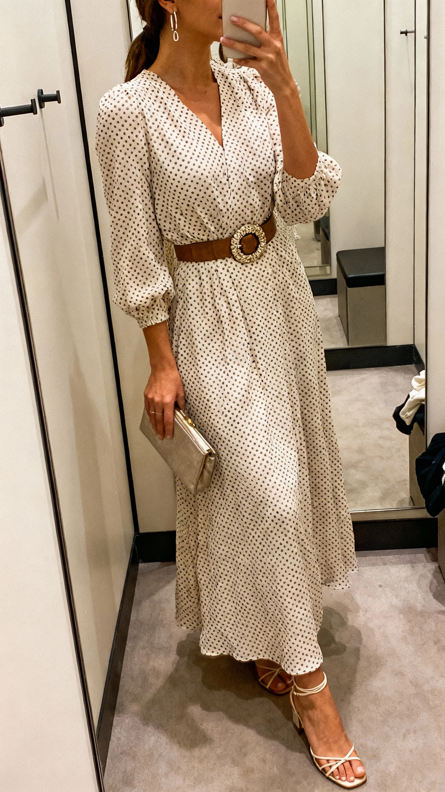 iPhone side-angle mirror selfie of a woman wearing a polka dot maxi dress cinched with a statement waist belt, delicate strappy heels, dainty earrings, and a small clutch, face not visible, cropped at shoulders, dressing-room mirror, warm indoor lighting, iPhone photo quality.