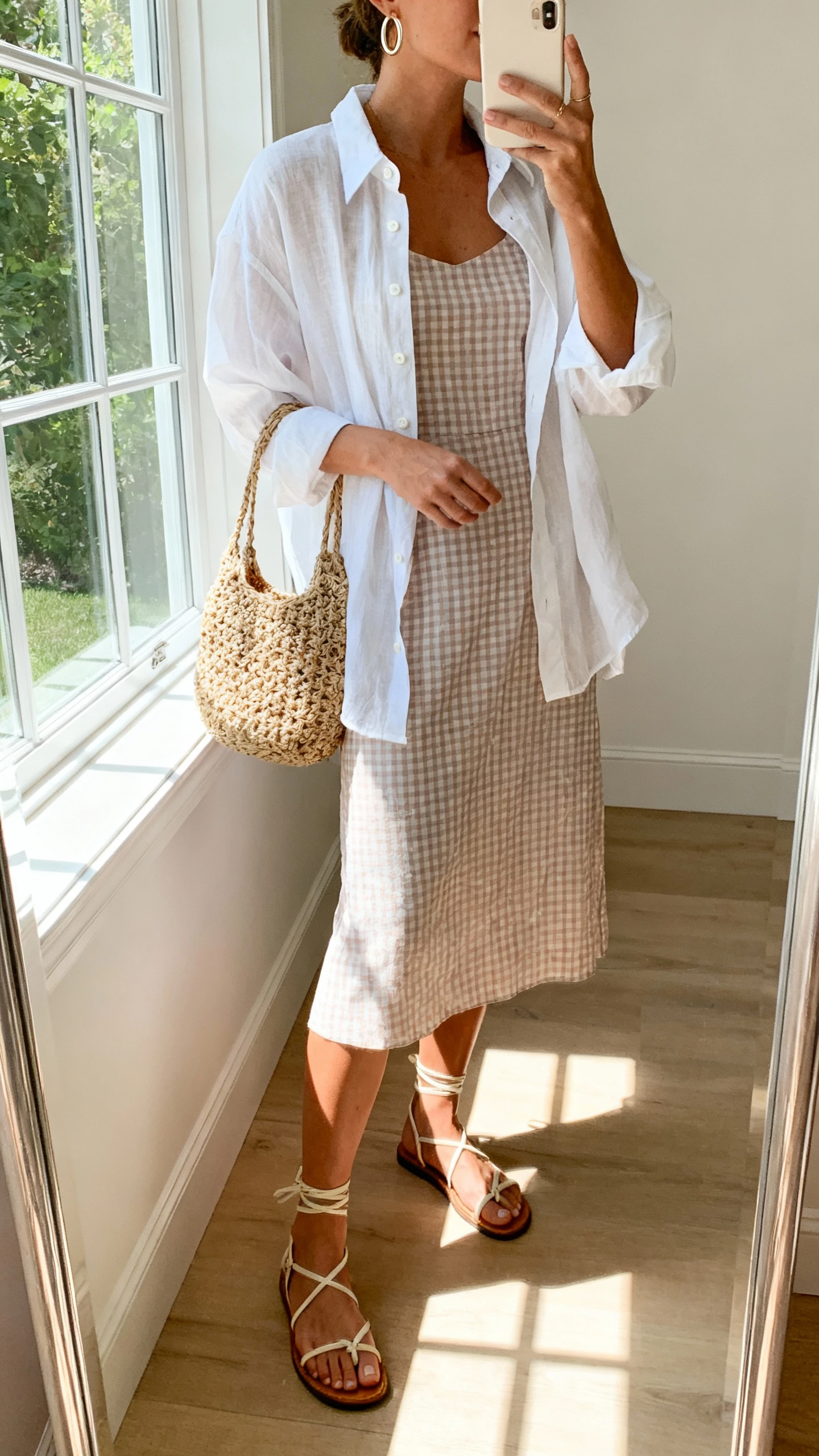 iPhone side-angle mirror selfie of a woman wearing a soft check sundress with an open white button-down as a breezy layer, strappy sandals, woven tote, and simple hoop earrings, face not visible, cropped at neck, natural daylight near window, iPhone photo quality.