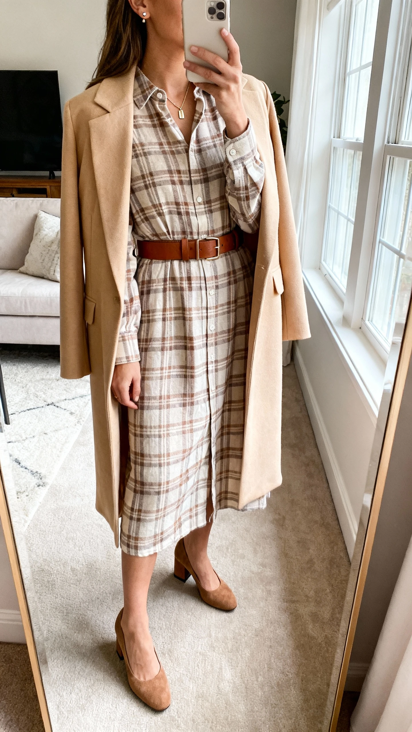 iPhone side-angle mirror selfie of a woman wearing a tonal plaid shirt dress in soft fabric (brushed cotton, Tencel, or lightweight flannel) with a chocolate or tan leather belt, soft suede pumps or rounded-toe block heels, stud earrings, a slim pendant/locket, and a tailored longline cardigan or waist-length blazer in cream or camel; face not visible, living room full-length mirror, morning window light, iPhone photo quality.
