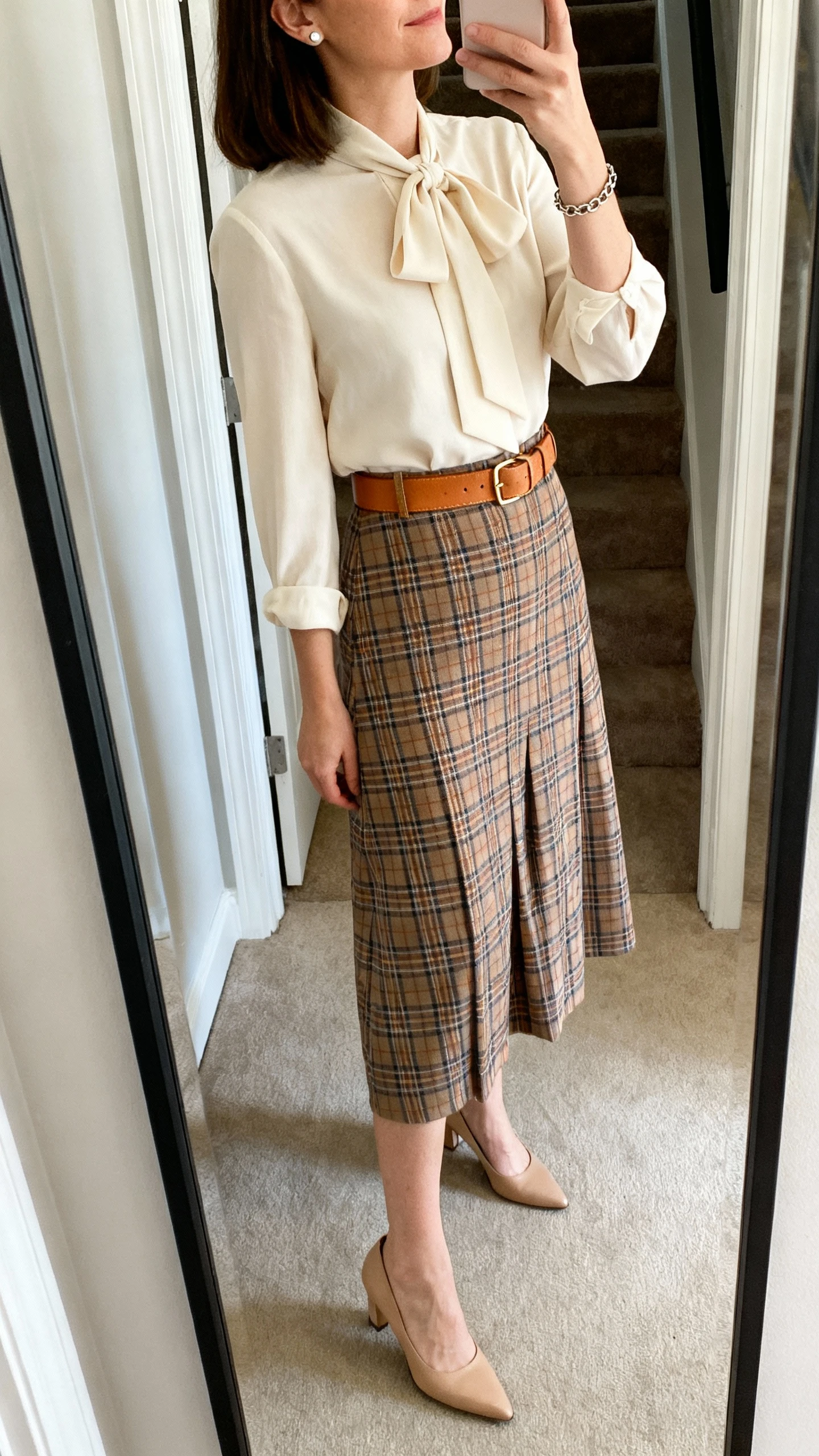 iPhone side-angle mirror selfie of a woman wearing a heritage plaid midi dress (or plaid midi skirt with a tan leather belt) paired with a cream bow-tie blouse, modest mid-heel pumps, stud earrings, and a simple chain bracelet, face not visible, cropped at neck, entryway full-length mirror, indoor daylight, iPhone photo quality.
