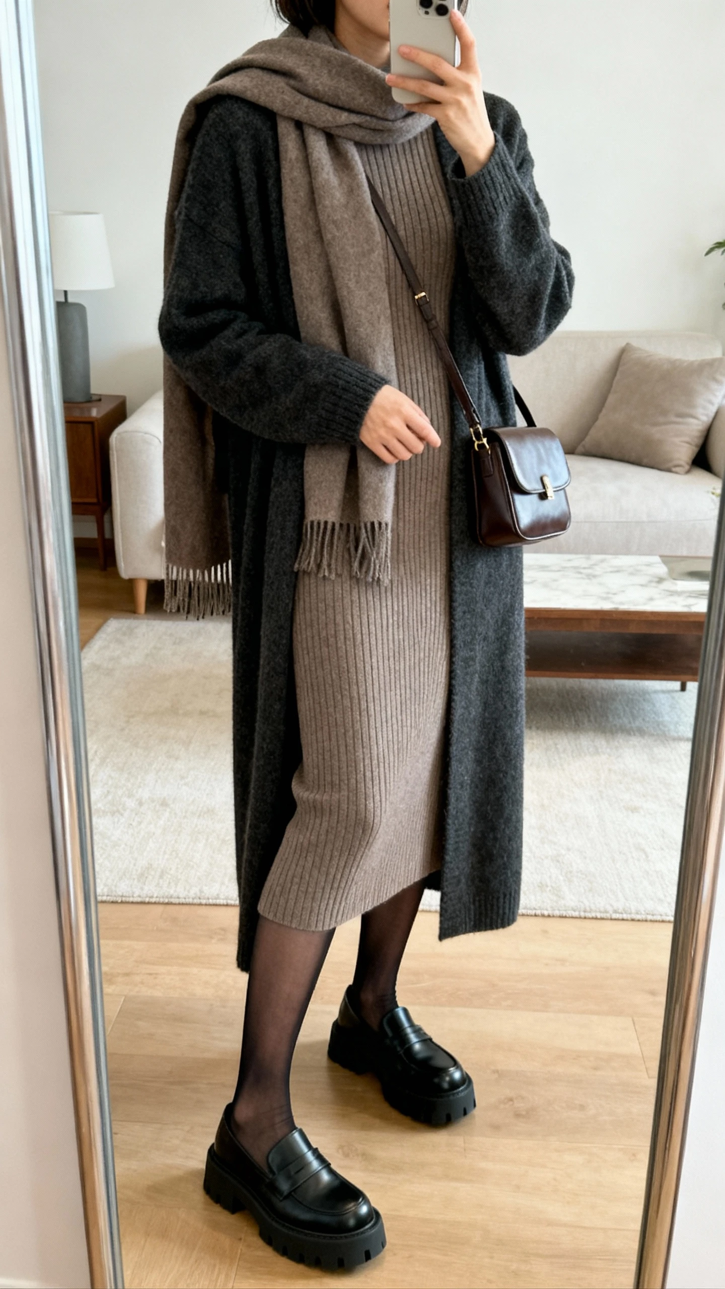 iPhone side-angle mirror selfie of a woman wearing a ribbed taupe knit midi dress layered with a long charcoal cardigan, sheer black tights, chunky black loafers, scarf draped over shoulders, small leather shoulder bag, face not visible, living room mirror, soft indoor lighting, iPhone photo quality.