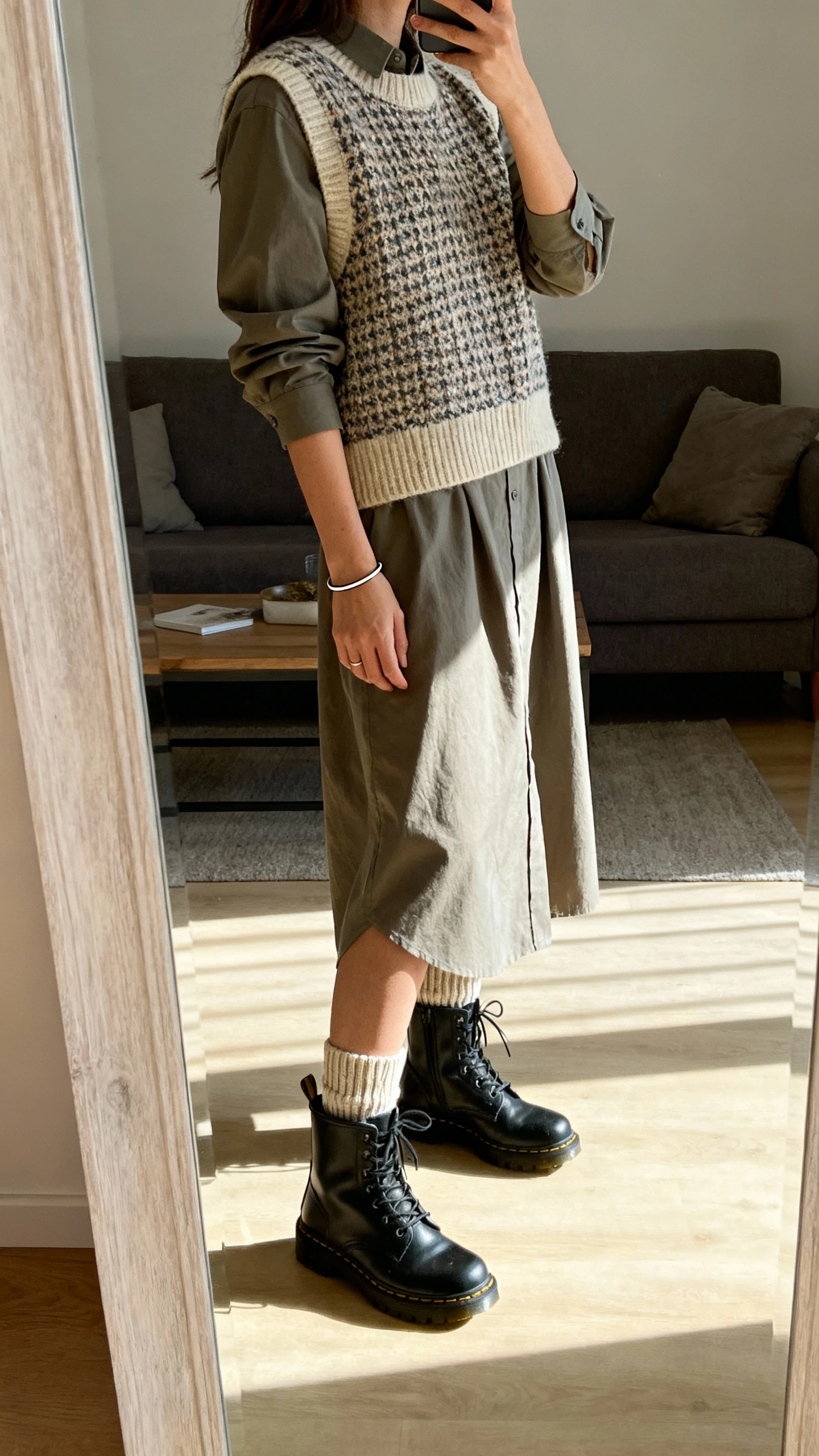 iPhone side-angle mirror selfie of a woman wearing a knit sweater vest layered over a long shirt dress with combat boots and ribbed socks, simple bracelet, face not visible, living room full-length mirror, afternoon natural light, iPhone photo quality.
