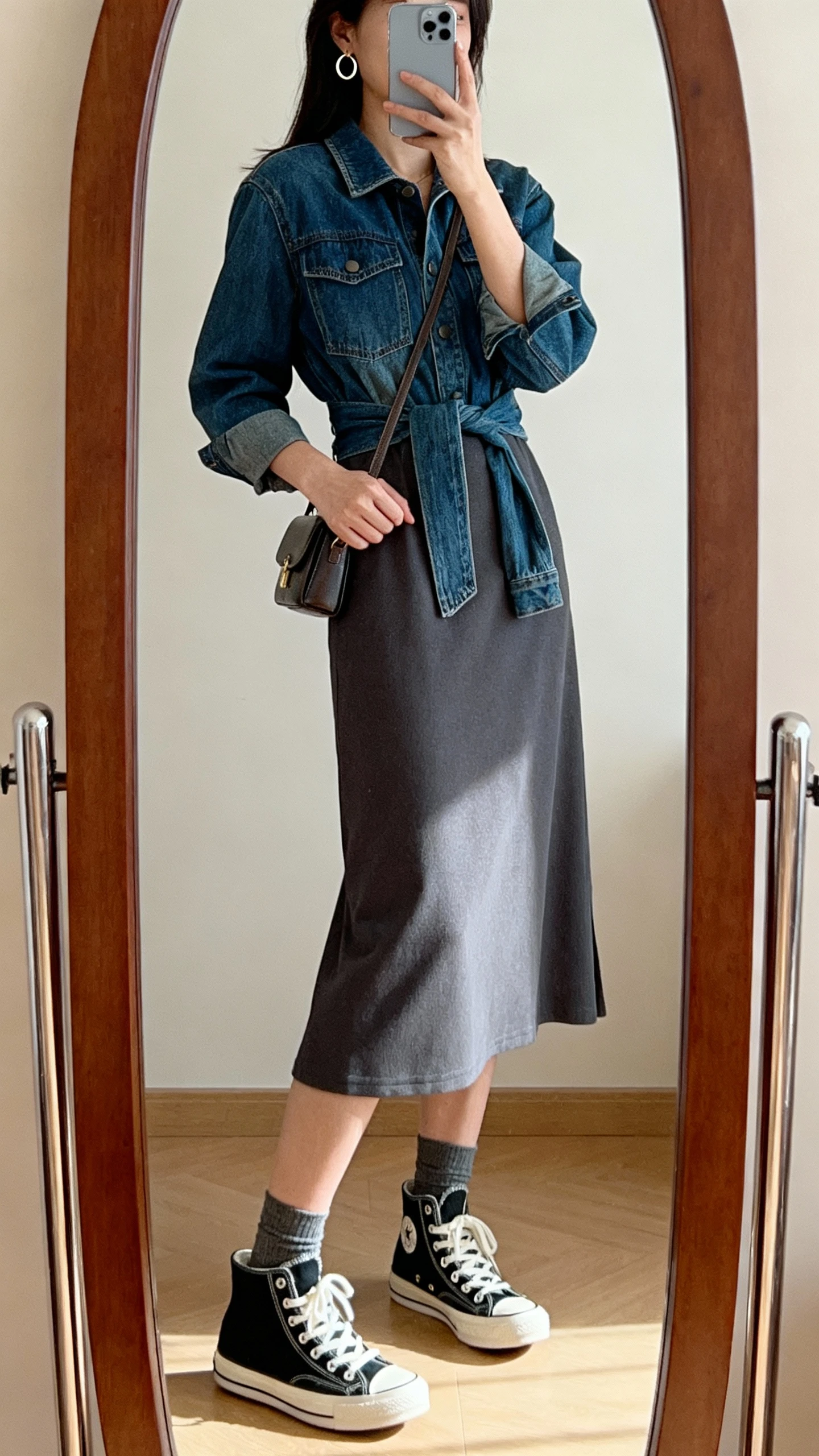 iPhone side-angle mirror selfie of a woman wearing a modest midi dress with high-top sneakers, ankle socks, denim jacket tied at the waist, small crossbody bag, and simple hoop earrings; face not visible, full-length mirror, warm indoor lighting, casual iPhone photo.