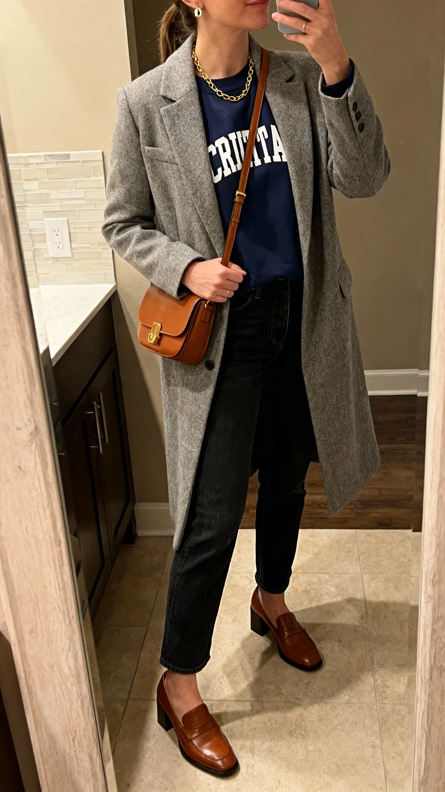 iPhone side-angle mirror selfie of a woman wearing a navy Christian crewneck under a heather gray longline blazer, dark straight-leg jeans, brown leather block-heel loafers, simple gold chain and stud earrings, and a cognac crossbody bag, face not visible, cropped at neck, warm indoor evening lighting, bathroom mirror, iPhone photo quality.