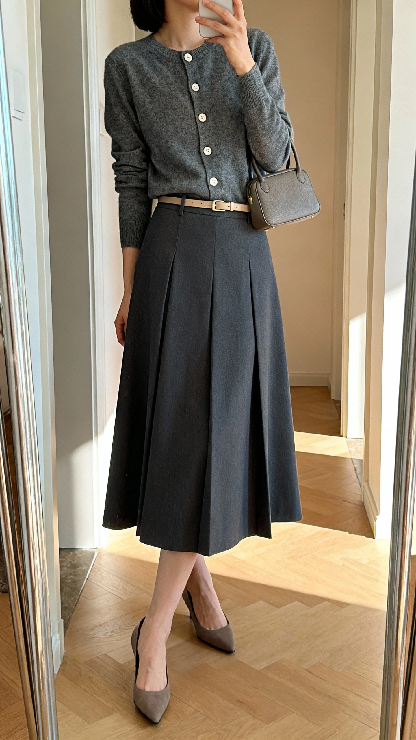 iPhone side-angle mirror selfie of a woman wearing a classic cardigan buttoned over an A-line skirt, low heels, thin belt, and a small top-handle bag, face not visible, cropped at neck, entryway full-length mirror, natural morning light, iPhone photo quality.