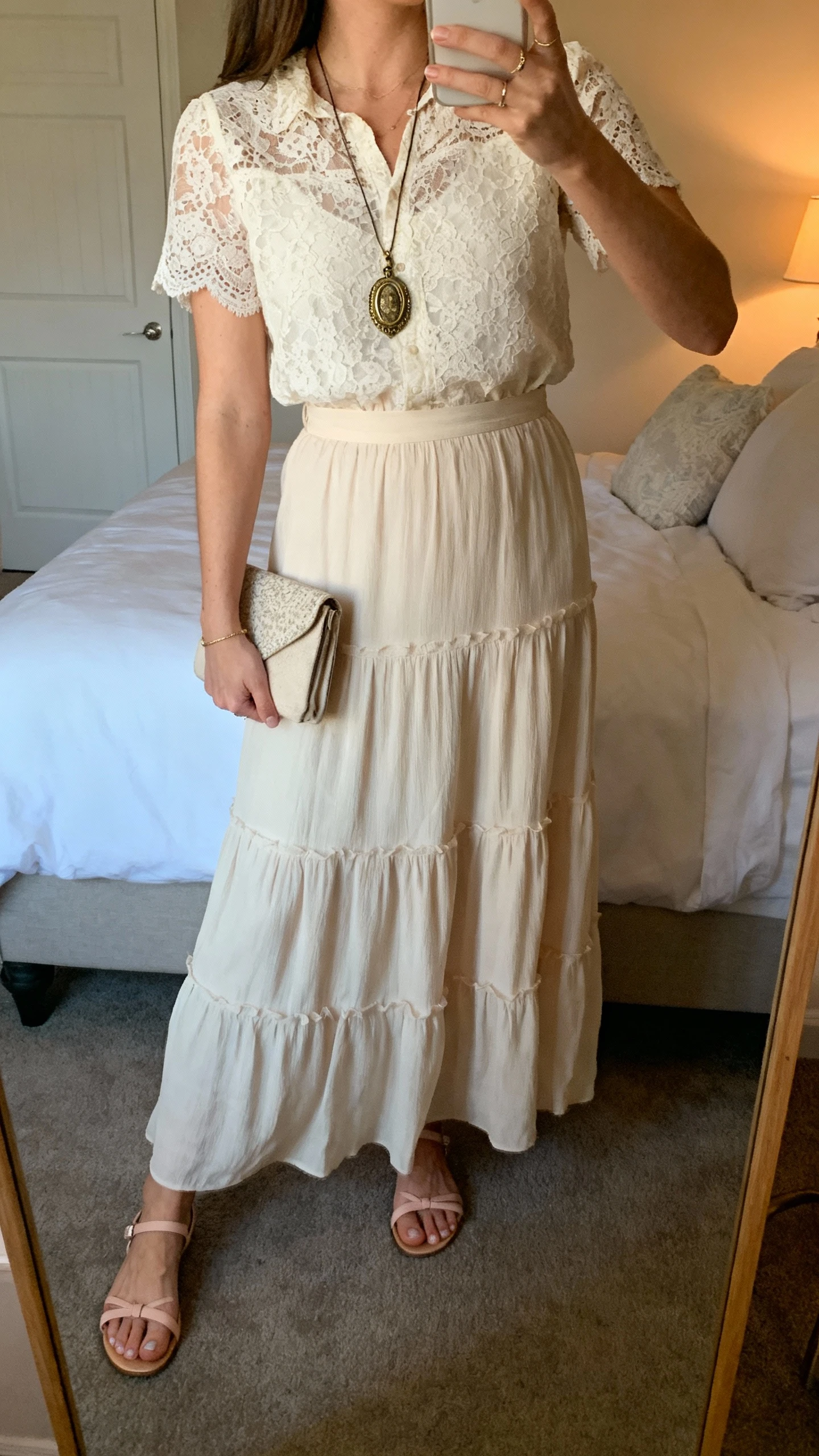 iPhone side-angle mirror selfie of a woman wearing a romantic lace top tucked into a tiered maxi skirt, vintage-inspired pendant necklace, small clutch, and dainty sandals, face not visible, bedroom mirror, soft evening indoor lighting, iPhone photo quality.