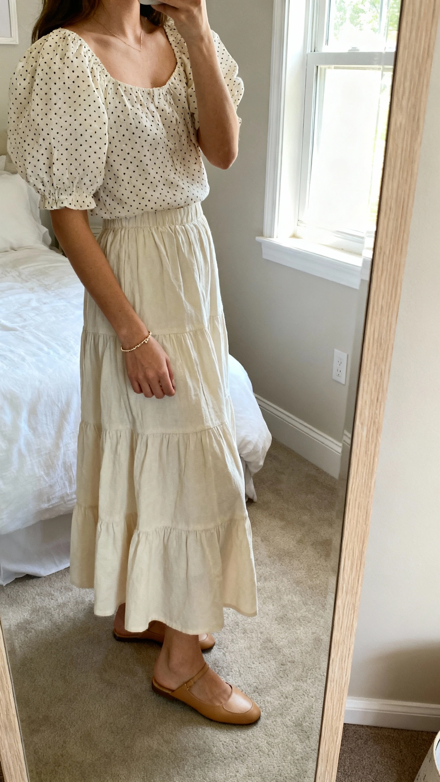iPhone side-angle mirror shot of a woman wearing a puff-sleeve Swiss dot blouse with a tiered cotton maxi skirt, dainty bracelet and flats, face not visible, cropped at shoulders, bedroom mirror, soft window light, iPhone photo quality.