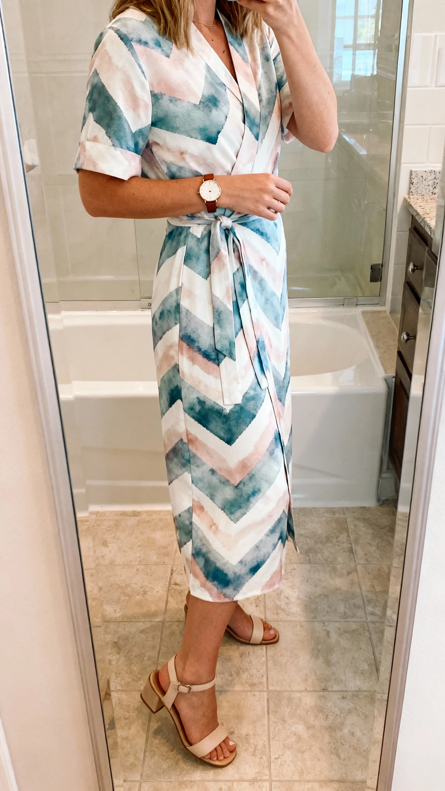 Side-angle iPhone mirror selfie of a woman in a watercolor chevron wrap dress that looks tailored but feels easy, with a slim leather watch and simple heeled sandals, face not visible, bathroom full-length mirror, natural indoor lighting, iPhone photo quality.