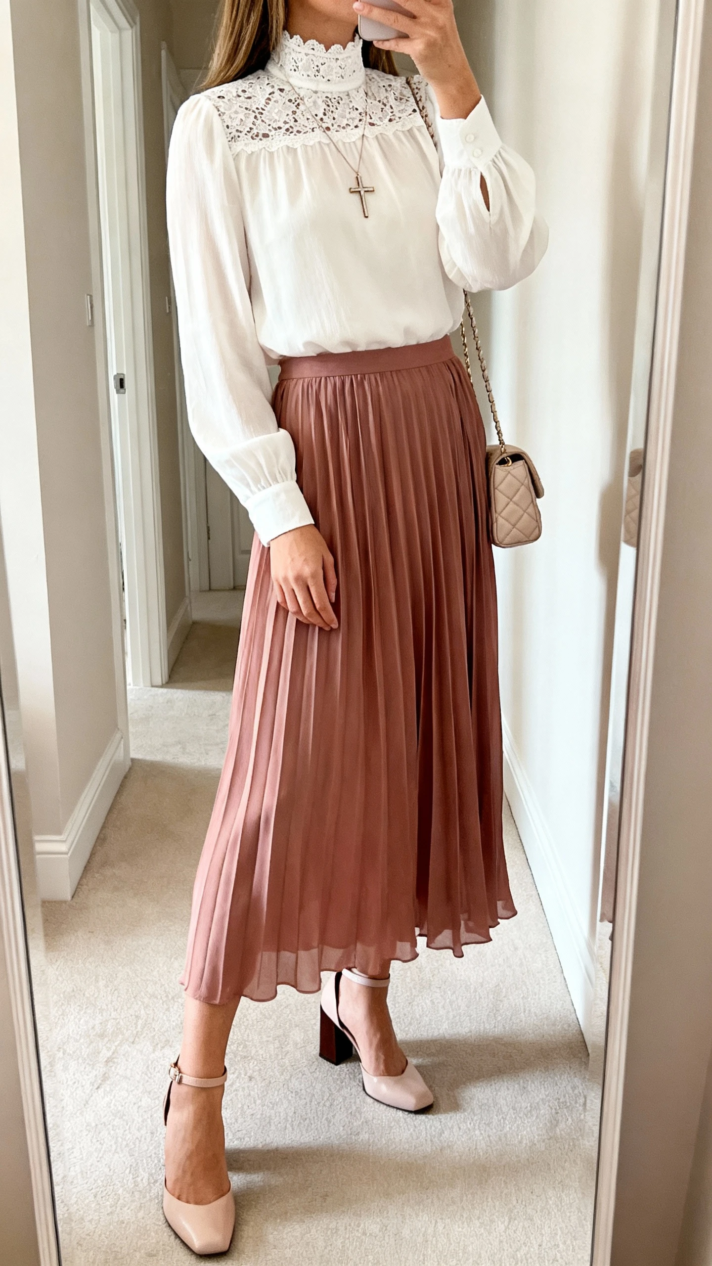 Side-angle iPhone mirror selfie of a woman wearing a romantic lace-trim high-neck blouse in ivory paired with a flowing dusty-rose pleated midi skirt, ankle-strap block heels, dainty cross pendant necklace, and a small quilted shoulder bag, face not visible, cropped at shoulders, full-length mirror in hallway, soft daylight, iPhone photo quality