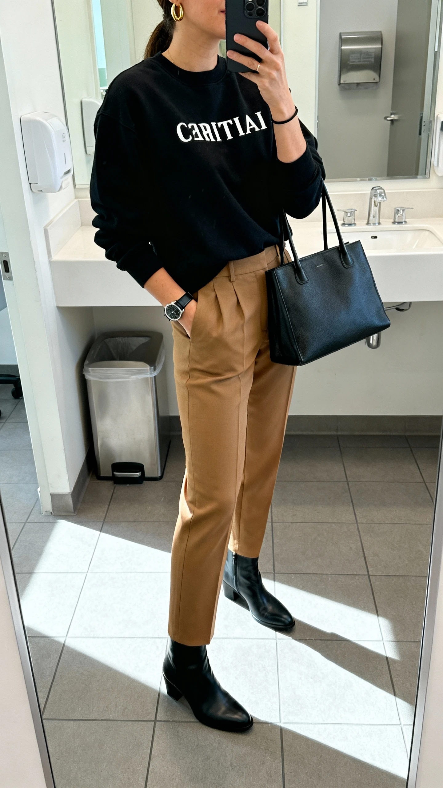 Side-angle iPhone mirror selfie of a woman wearing a black Christian crewneck tucked into tailored camel trousers, sleek black ankle boots, minimalist gold hoops, thin black leather watch, and a structured black tote, face not visible, cropped at shoulders, office bathroom mirror, bright natural light, iPhone photo quality.