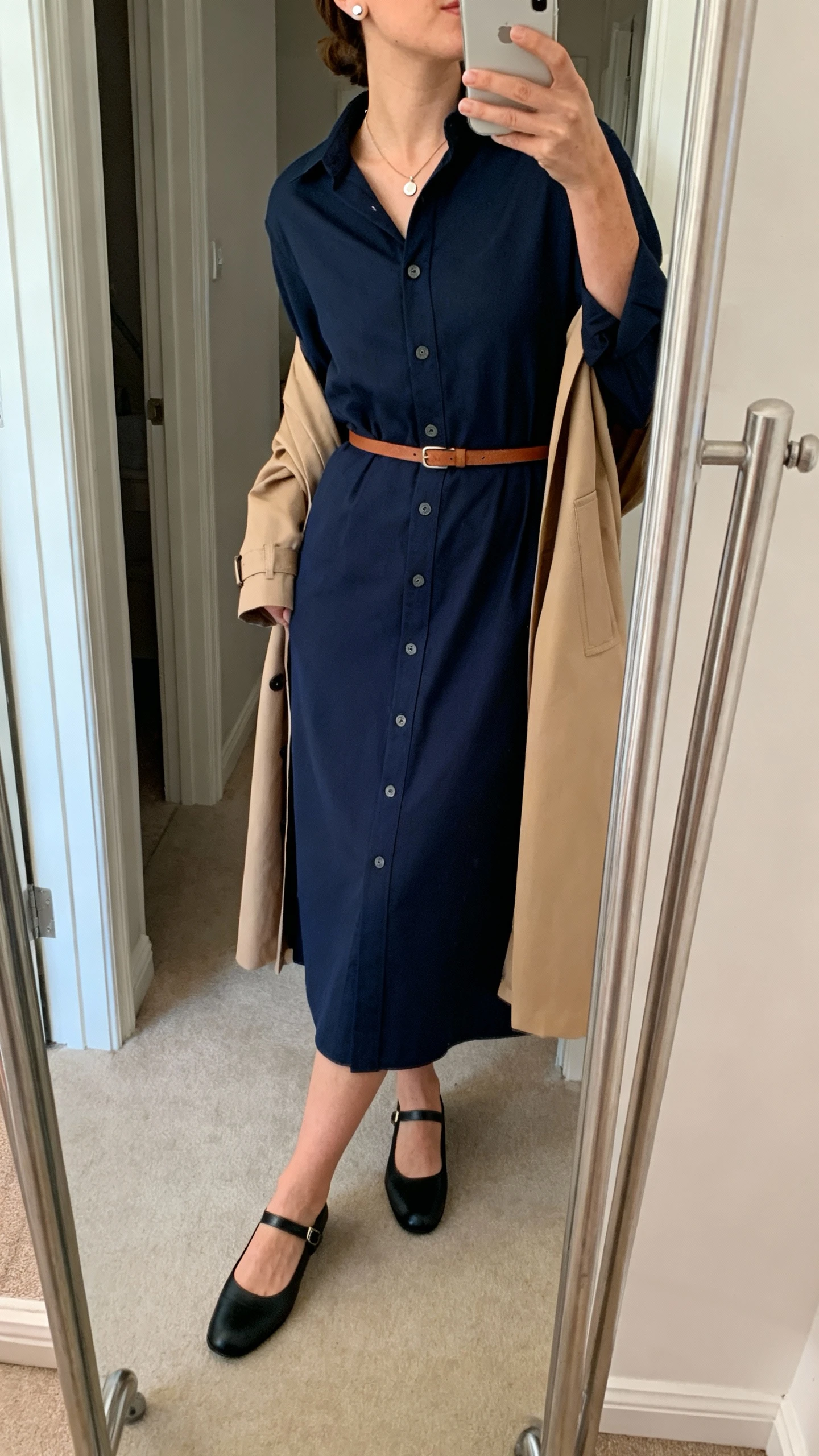 Side-angle iPhone mirror selfie of a woman wearing a navy button-front midi shirt dress with a thin leather belt, sleek black Mary Janes, a light tan trench coat draped over shoulders, simple pendant and stud earrings, face not visible, cropped at neck, entryway full-length mirror, natural daylight, iPhone photo quality.