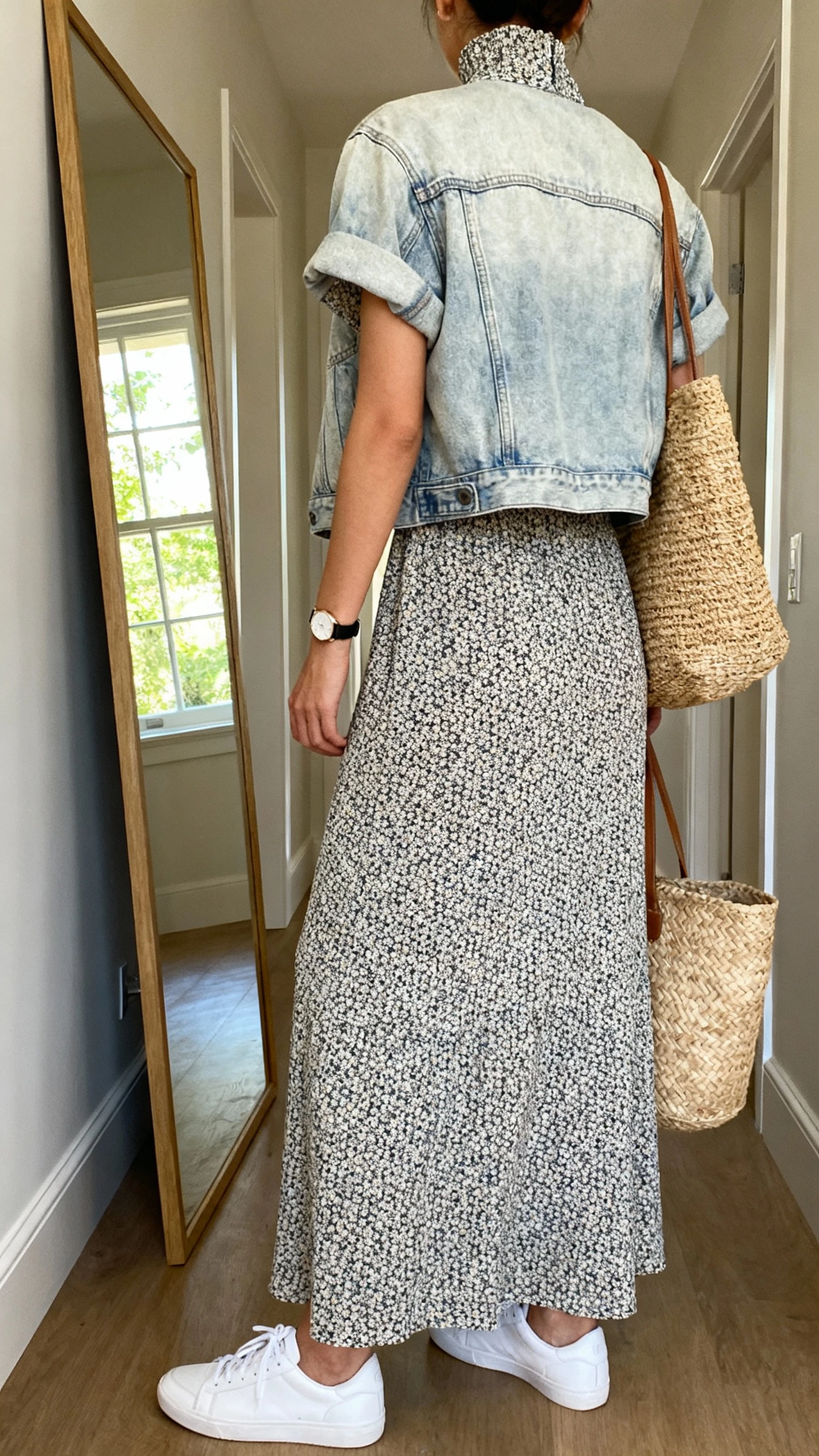 iPhone back view of a woman wearing a micro-print maxi dress with a high neckline and short sleeves, light-wash slightly boxy denim jacket, white sneakers, woven straw tote, simple watch; face not visible, hallway full-length mirror, natural daylight from window, iPhone photo quality.
