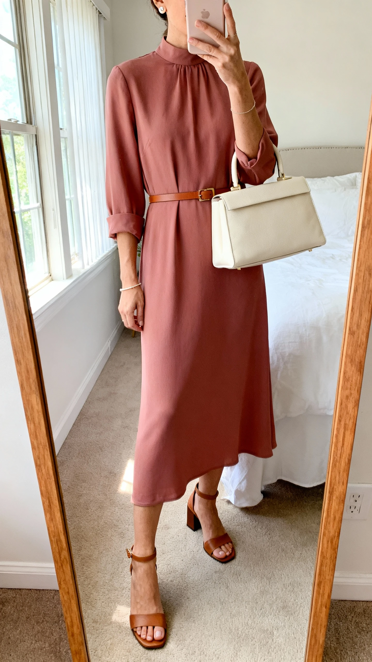 iPhone mirror selfie of a woman wearing a solid midi dress with a high neckline and elbow sleeves in dusty rose, thin cognac waist belt, low block-heel sandals, structured cream top-handle bag, minimal stud earrings, and a delicate bracelet; face not visible, cropped at neck, bedroom mirror, natural morning window light, iPhone photo quality.