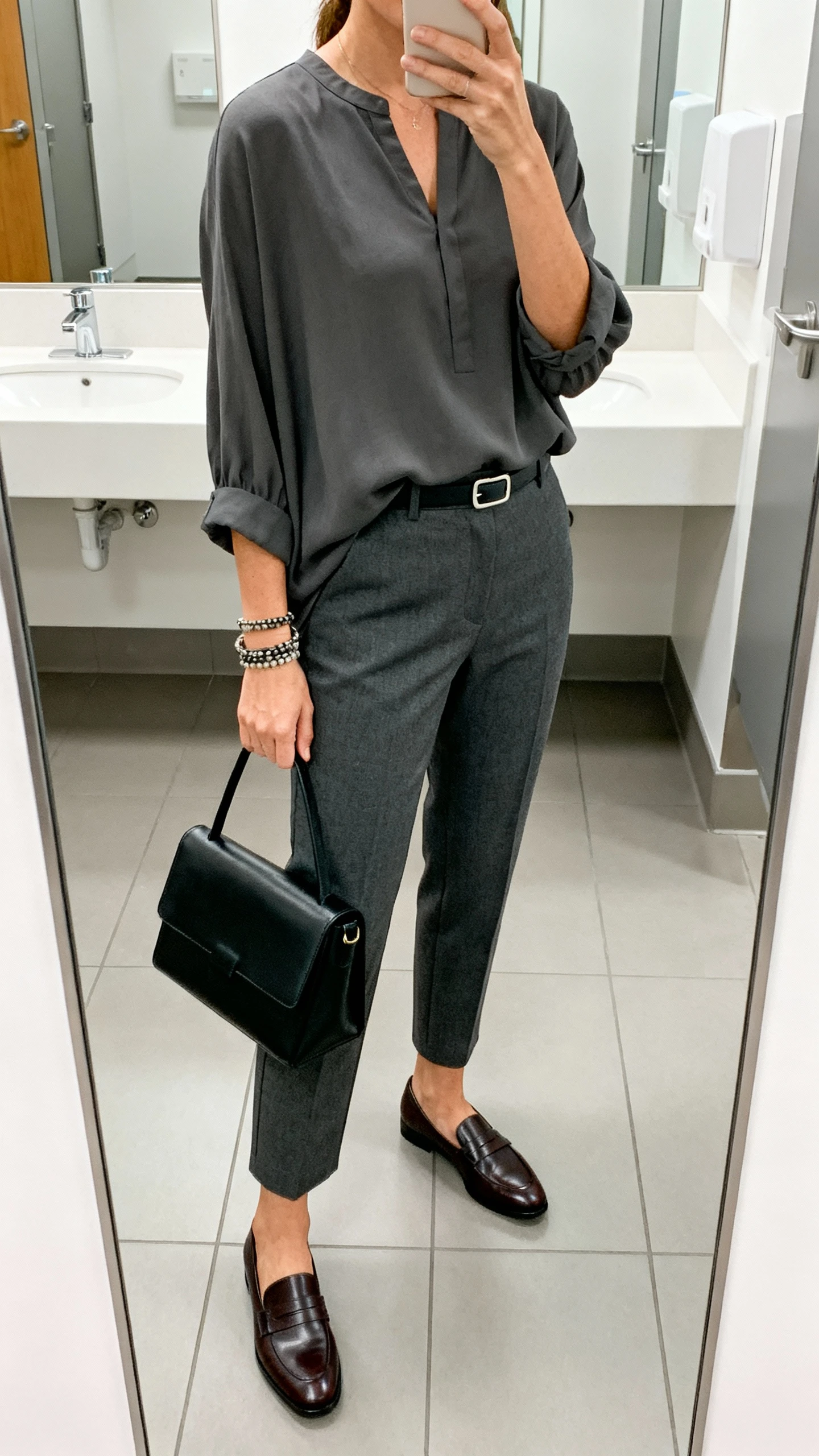 iPhone mirror selfie of a woman wearing high-waisted straight-leg trousers in charcoal, hip-length tunic blouse with a modest neckline and bracelet sleeves, leather loafers, minimalist belt, structured black satchel; face not visible, cropped at shoulders, office bathroom mirror, bright indoor lighting, iPhone photo quality.