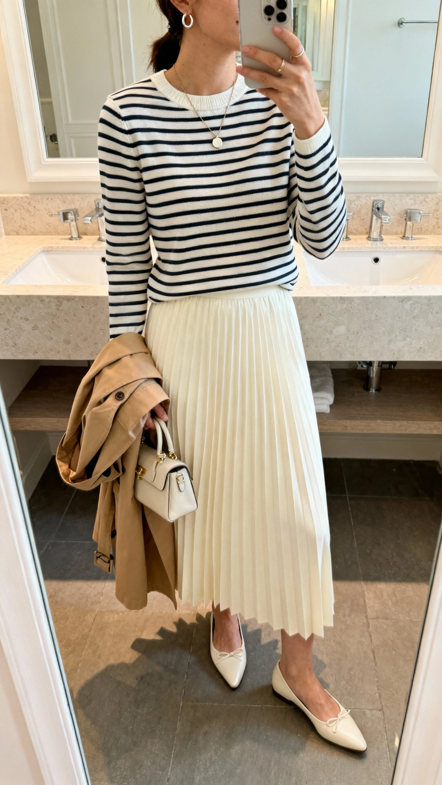 iPhone side-angle mirror selfie of a woman wearing a cream midi pleated skirt, long-sleeve Breton stripe top with a modest crew neck, pointed ballet flats, lightweight camel trench draped over arm, simple pendant necklace and small hoop earrings, holding a small handbag; face not visible, bathroom mirror, soft indoor light, iPhone photo quality.