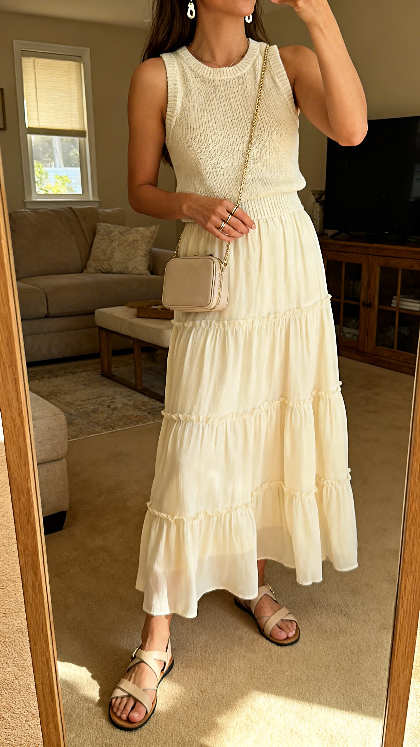 iPhone mirror selfie of a woman in a flowy tiered cream maxi skirt with a fine knit high-crew tank, strappy flat sandals, a compact crossbody, delicate drop earrings, and slim stacking rings; face not visible, cropped at shoulders, living room full-length mirror, warm indoor light, iPhone photo quality.