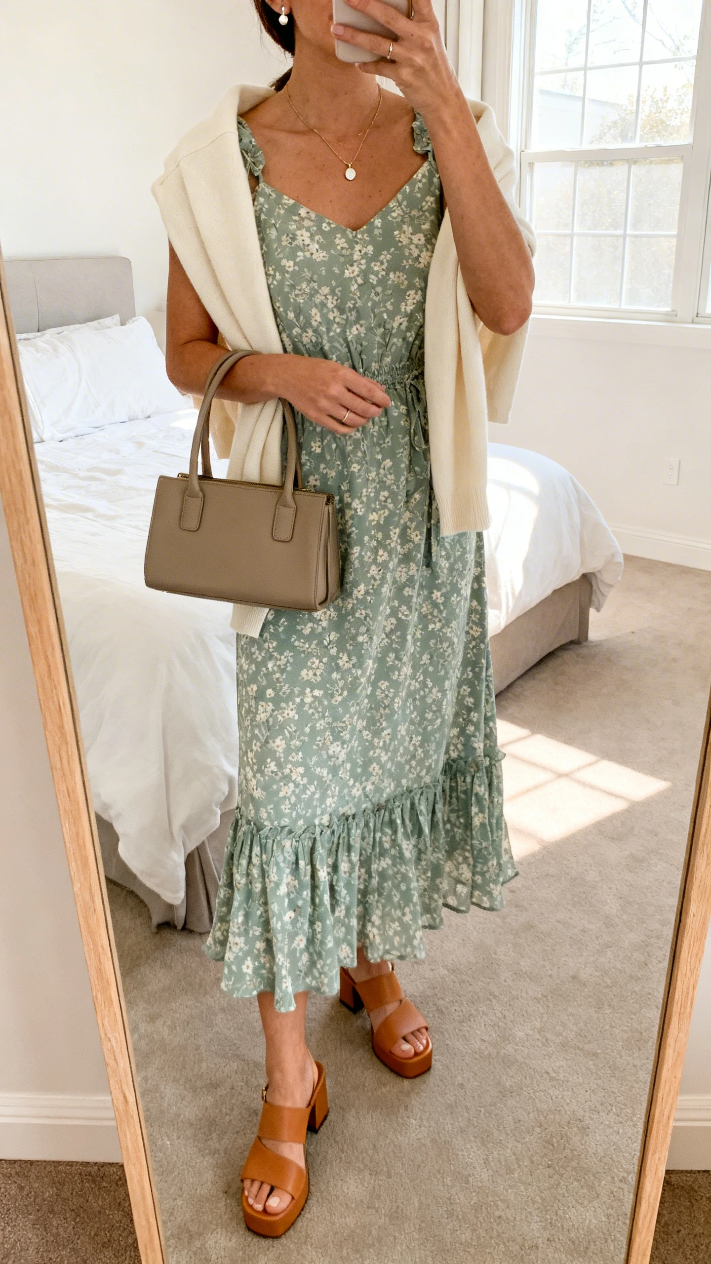iPhone mirror selfie of a woman wearing a breezy floral midi dress in sage with tan low block-heel sandals, a structured top-handle bag, delicate pendant necklace, simple stud earrings, and a light cream cardigan draped over her shoulders; face not visible, cropped at neck, bedroom mirror, soft morning window light, iPhone photo quality.